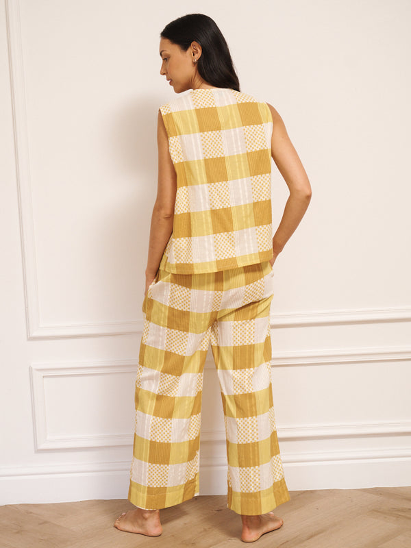 Model wearing a golden gingham cotton waistcoat set with a sleeveless button-front top and wide-leg trousers, standing barefoot against a white studio wall with a light wooden floor. Back detail