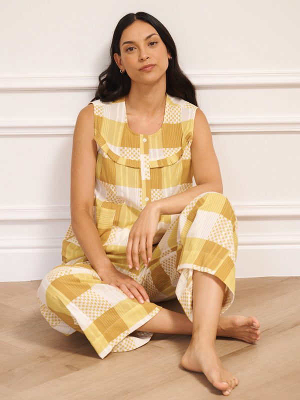 Model wearing a golden gingham cotton waistcoat set with a sleeveless button-front top and wide-leg trousers, standing barefoot against a white studio wall with a light wooden floor. Sat on floor