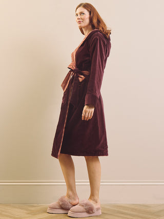 A woman wearing a plush burgundy velvet dressing gown robe with a tie waist, standing in a room with a beige wall and light wood floor, wearing pink slippers.