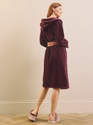 A woman wearing a plush hooded dressing gown robe in deep burgundy, shown from the back and side. She is wearing light pink slippers in a minimalist room with beige walls and a light wood floor.