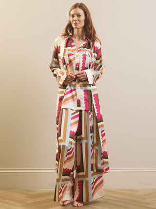 A woman stands barefoot in a studio wearing a matching long-sleeve button-up shirt and wide-leg pants set with a bold geometric pattern in beige, brown, pink, and white.
