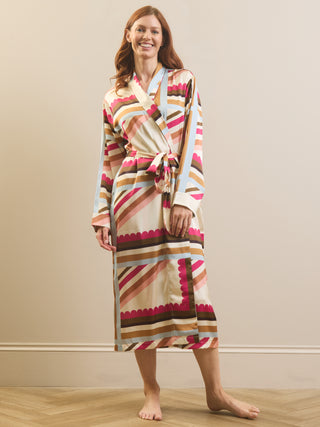A woman modelling a long-sleeve wrap dressing gown robe with a colourful geometric stripe pattern in pink, brown, beige, and light blue, standing against a plain beige wall.