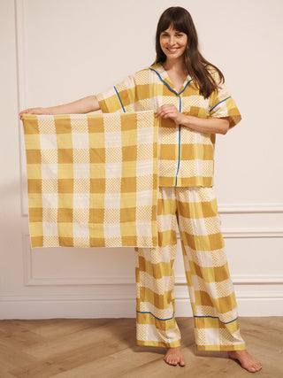 Woman wearing yellow checkered pyjamas holding a matching scarf in a room with wooden flooring and white walls.