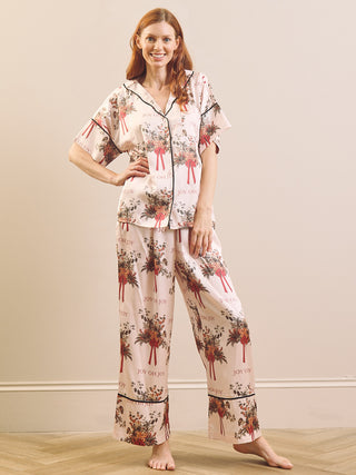 A woman modelling a two-piece pyjama set with a floral print and black piping, standing against a neutral beige wall and light wood floor.