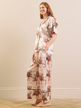 A woman modelling a two-piece pyjama set with a floral and ribbon print on a light beige background, standing against a plain beige wall on a light wood floor.