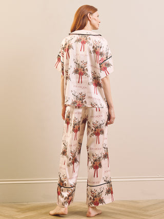 A woman stands barefoot indoors wearing a matching pyjama set with a light pink base and an allover floral bouquet print with red ribbon accents and black piping.