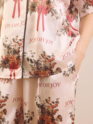 A close-up of a person wearing a matching pajama set with a cream base, red 'JOY OH JOY' text, and red ribbon motifs over a floral print.