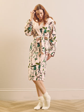 A woman wearing a long-sleeve hooded dressing gown robe with a botanical pear print in green, brown, and beige on a light pinkish background, paired with white socks, standing in a room with a beige wall and light wood floor.