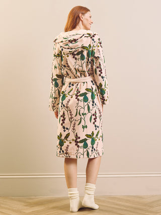 A woman stands with her back to the camera wearing a hooded long-sleeve velvet robe with an pear branch botanical print. The robe has a light pinkish-beige base with green leaves and dark olive-brown olives, paired with cream-colored socks.