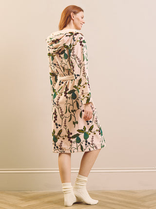 A woman stands in profile wearing a hooded velvet robe with a botanical pear print. The robe has a light blush pink base with green leaves and dark berry motifs, and is styled with cream socks against a neutral beige wall and light wood floor.