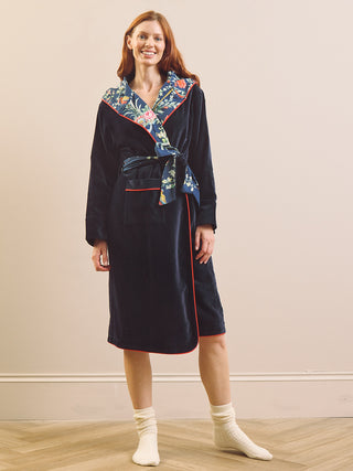 A woman wearing a dark navy velvet bathrobe with a colorful floral patterned collar and red piping, standing in a studio with a beige wall and light wood floor.