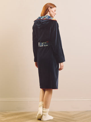 A woman wearing a dark navy velvet hooded robe with a colorful floral patterned inner hood lining and a matching patterned waist tie, shown from the back against a beige wall.