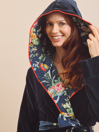 A smiling woman wearing a dark navy velvet hooded dressing gown robe with a colourful floral and bird print lining and red piping, posed against a plain beige background.