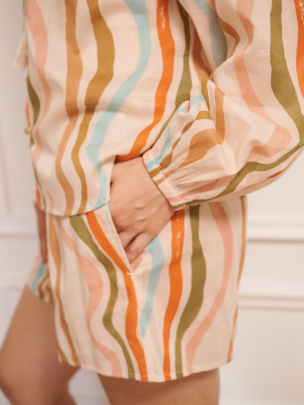 Model wearing women’s cotton voile short pyjama set in oatmeal with rainbow wave print, featuring long-sleeve raglan top with tie neckline and matching short pyjama bottoms.