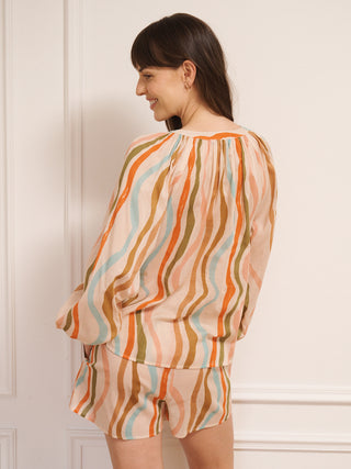 Model wearing women’s cotton voile short pyjama set in oatmeal with rainbow wave print, featuring long-sleeve raglan top with tie neckline and matching short pyjama bottoms.