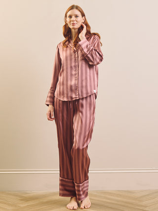A woman modelling a two-piece pyjama set with vertical stripes in dusty rose and brown, standing against a plain beige wall on a light wood floor.