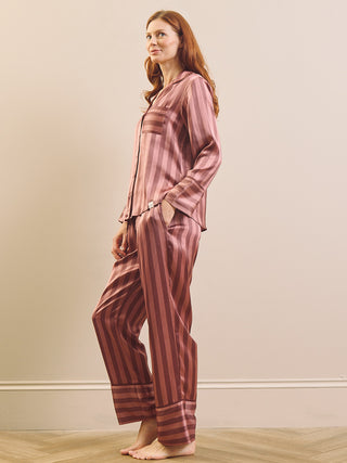 A woman modelling a matching two-piece pyjama set with vertical stripes in dusty rose and dark brown, standing barefoot against a neutral beige wall.