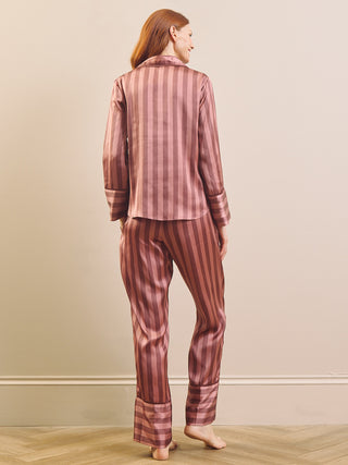 A woman stands barefoot indoors wearing a matching two-piece pyjama set with vertical stripes in dusty rose and dark brown. The set includes a long-sleeve button-up top and coordinating pants, shown from the back against a plain beige wall and light wood floor.