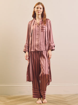 A woman modeling a matching two-piece pajama set with a long-sleeve button-front top and wide-leg pants and matching robe, both in a dusty rose and brown vertical stripe pattern, against a plain beige wall.