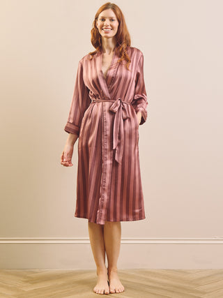 A woman wearing a dusty rose pink satin robe with vertical stripes, standing barefoot in a room with a beige wall and light wood floor.