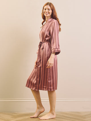 A woman modeling a satin-like robe with vertical stripes in dusty rose and dark brown, standing barefoot against a plain beige wall with a light wood floor.