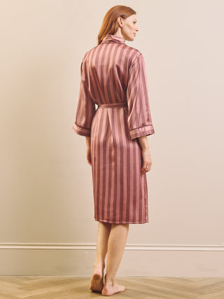 A woman stands barefoot in profile wearing a satin-like robe with vertical pink and dusty rose stripes, photographed against a plain beige wall and light wood floor.