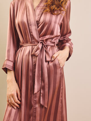 A woman wearing a long-sleeve satin robe with vertical dusty rose and mauve stripes, tied at the waist with a matching belt, against a plain beige background.