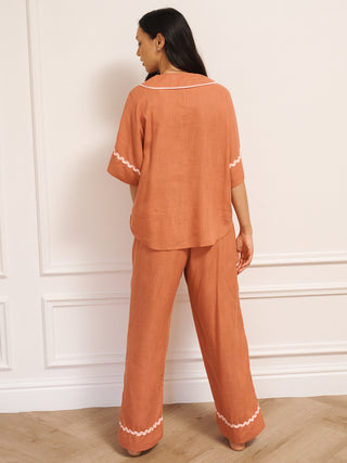 Model wearing women’s oversized linen blend pyjamas in rust with pale pink contrast trim, featuring short-sleeve button-front top and wide-leg trousers in a relaxed fit. Back Detail