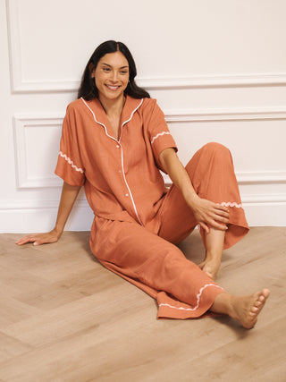 Model wearing women’s oversized linen blend pyjamas in rust with pale pink contrast trim, featuring short-sleeve button-front top and wide-leg trousers in a relaxed fit. Sat on the floor