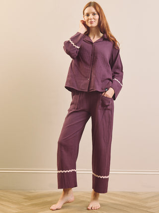 A woman modelling a matching two-piece pyjama set in a muted plum purple colour. The set includes a long-sleeve button-up shirt and matching pants, both made of a textured, vertically ribbed fabric, with delicate white lace trim detailing the cuffs and hems.