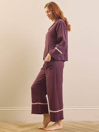 A woman modelling a matching two-piece pyjama set in deep plum purple, featuring a long-sleeve button-front top with decorative white lace trim on the cuffs and matching wide-leg pants with lace trim at the hems, set against a neutral beige wall and light wood floor.
