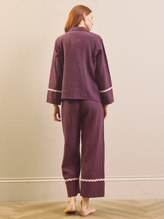 A woman stands barefoot indoors wearing a matching mauve-purple pyjama set with white zigzag trim on the cuffs and hems, photographed from the back against a plain beige wall.