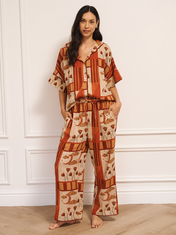 Model wearing women’s oversized linen blend pyjamas with natural patchwork sky flower print, featuring short-sleeve button-front shirt and wide-leg trousers in warm rust and neutral tones.