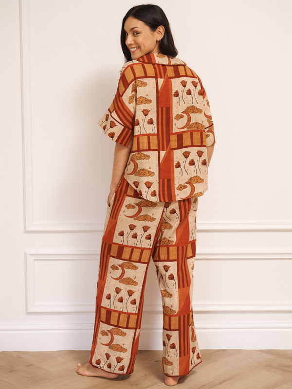 Model wearing women’s oversized linen blend pyjamas with natural patchwork sky flower print, featuring short-sleeve button-front shirt and wide-leg trousers in warm rust and neutral tones.