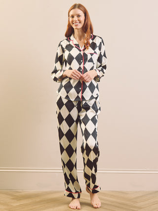 A woman modeling a two-piece pyjama set with a black and cream harlequin diamond pattern, featuring a button-front top with red piping and matching patterned pants, set against a neutral beige wall and light wood floor.