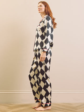 A woman stands barefoot in profile wearing a matching long-sleeve pyjama set with a black and cream harlequin diamond pattern and pink piping accents.