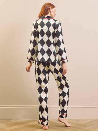 A woman stands barefoot against a beige wall, viewed from the back, wearing a matching long-sleeve pyjama set with a black and cream harlequin diamond pattern and thin pink piping along the collar, cuffs, and hem.