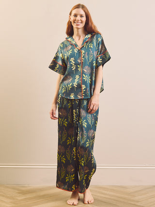 A woman modelling a matching two-piece pyjama set with a dark emerald green background and a botanical pattern in yellow, green, and brown, featuring a short-sleeve button-up top and long pants.
