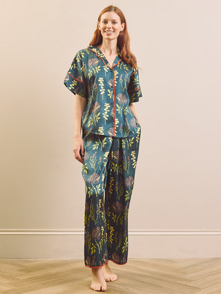 A woman modelling a matching short-sleeve button-front pyjama top and wide-leg pants set in a dark teal-blue color with a botanical leaf pattern in yellow, green, and orange, accented with red piping.