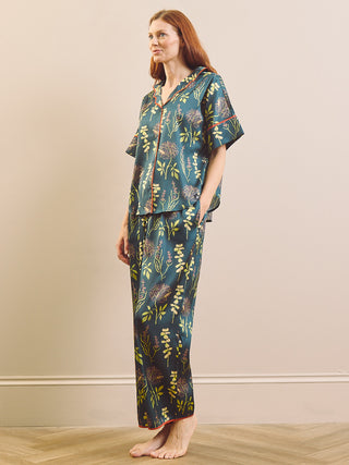 A woman stands barefoot in a studio wearing a matching short-sleeve button-front top and wide-leg pants pyjama set in a dark teal-blue colour with a botanical print in green, yellow, and orange.