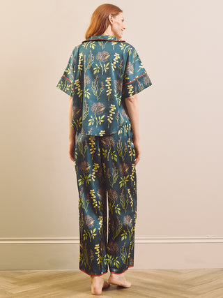 A woman stands barefoot indoors wearing a matching short-sleeve top and wide-leg pants pyjama set in a dark teal green color with a yellow and brown botanical pattern.