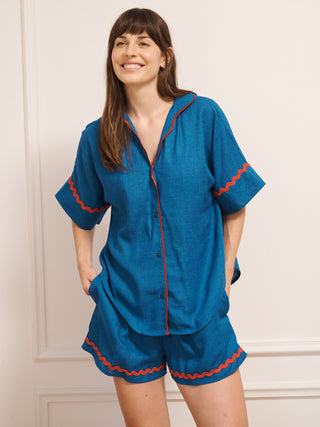 Model wearing women’s oversized linen blend short pyjama set in cobalt blue with red contrast ric-rac trim, featuring short-sleeve button-front shirt and matching elasticated shorts.