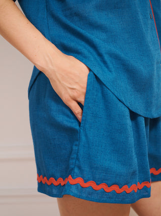 Model wearing women’s oversized linen blend short pyjama set in cobalt blue with red contrast ric-rac trim, featuring short-sleeve button-front shirt and matching elasticated shorts. Pocket detail