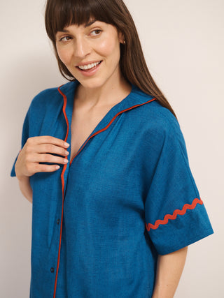 Model wearing women’s oversized linen blend short pyjama set in cobalt blue with red contrast ric-rac trim, featuring short-sleeve button-front shirt and matching elasticated shorts. Close up of top