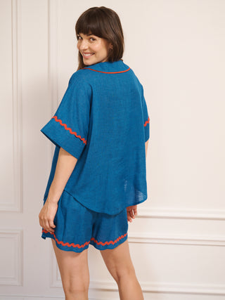 Model wearing women’s oversized linen blend short pyjama set in cobalt blue with red contrast ric-rac trim, featuring short-sleeve button-front shirt and matching elasticated shorts. Back detail