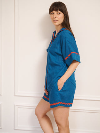 Model wearing women’s oversized linen blend short pyjama set in cobalt blue with red contrast ric-rac trim, featuring short-sleeve button-front shirt and matching elasticated shorts.