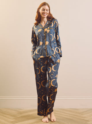 A woman stands barefoot in a bedroom setting wearing a matching pyjama set with a dark blue base and a repeating celestial print of gold crescent moons,  stars and cats.