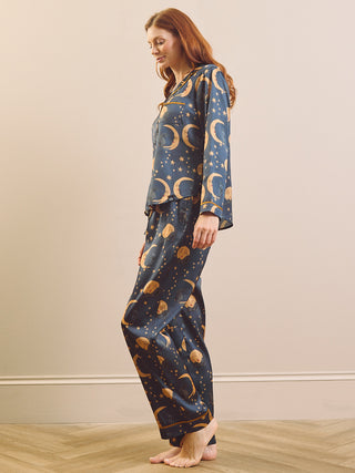 A woman modelling a two-piece pyjama set with a dark blue space-themed print featuring gold crescent moons, stars, and planets, standing against a beige wall and light wood floor.