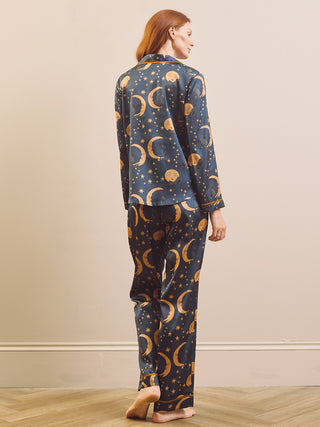 A woman stands barefoot indoors wearing a matching pyjama set with a dark blue background and an allover celestial print of gold crescent moons, stars, and planets, shown from the back.