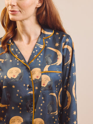 A close-up of a woman wearing navy blue satin pyjamas with a celestial print of gold crescent moons, stars, and cats, outlined with gold piping.
